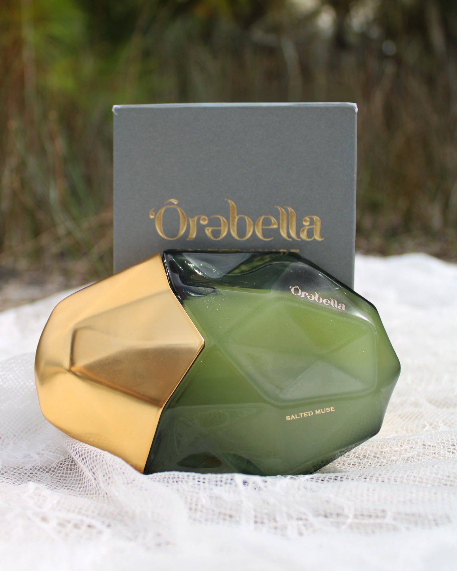 Salted Muse — the green faceted bottle, its grey outer box, on white linen in the marsh grass